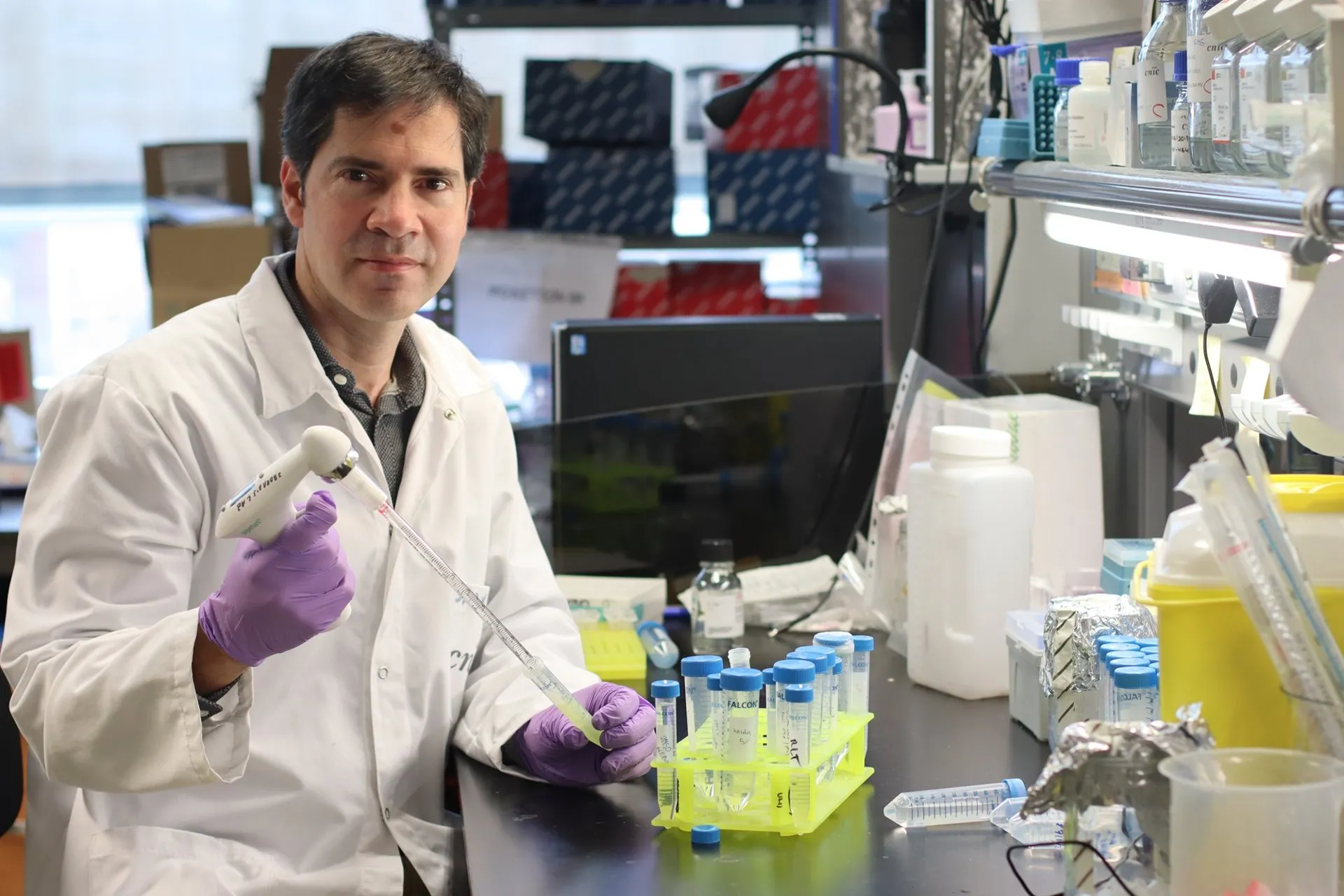 David Sancho, con bata blanca y guantes morados, sostiene una pipeta junto a una gradilla amarilla de tubos en su laboratorio