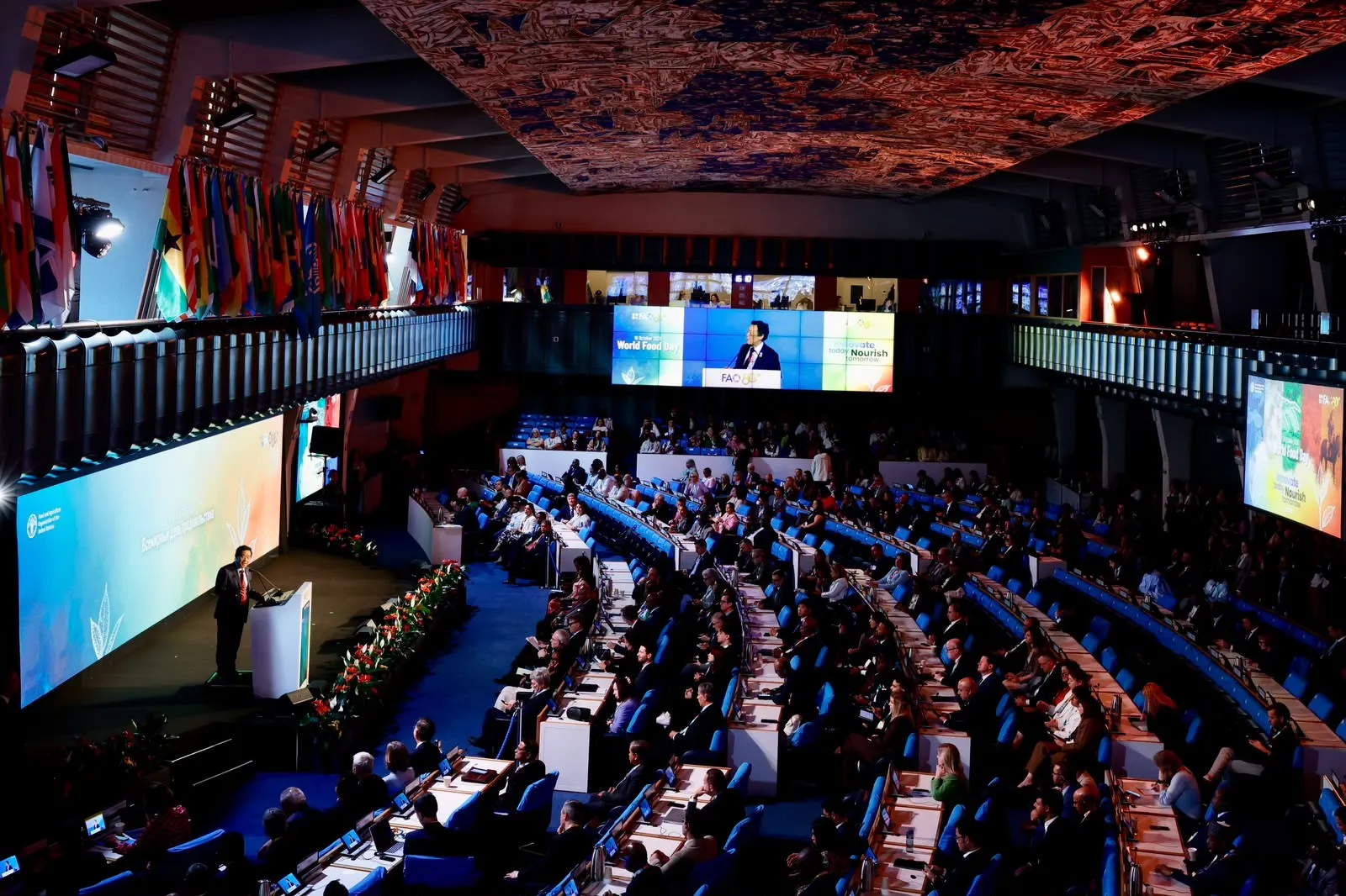 Gran sala de la FAO en Roma vista desde arriba, con banderas en el balcón, cientos de asistentes en filas de escaños azules y un orador en el atril junto a una pantalla gigante