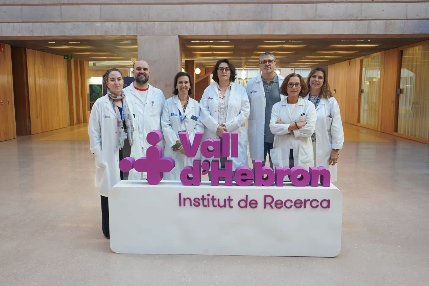 Siete profesionales con bata blanca posan de pie detrás del rótulo morado de Vall d'Hebron Institut de Recerca, en el vestíbulo del edificio