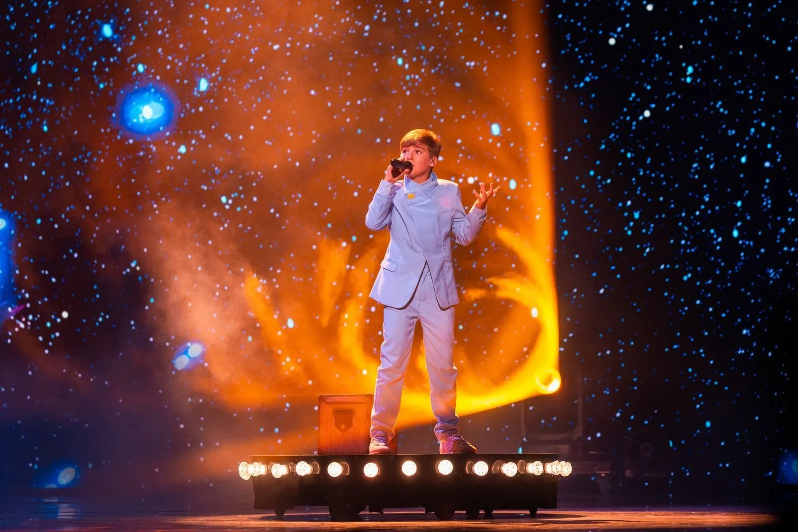 Lucas Paulano, de pie sobre una plataforma con focos, canta ante una pantalla con un cielo de estrellas y una estela naranja