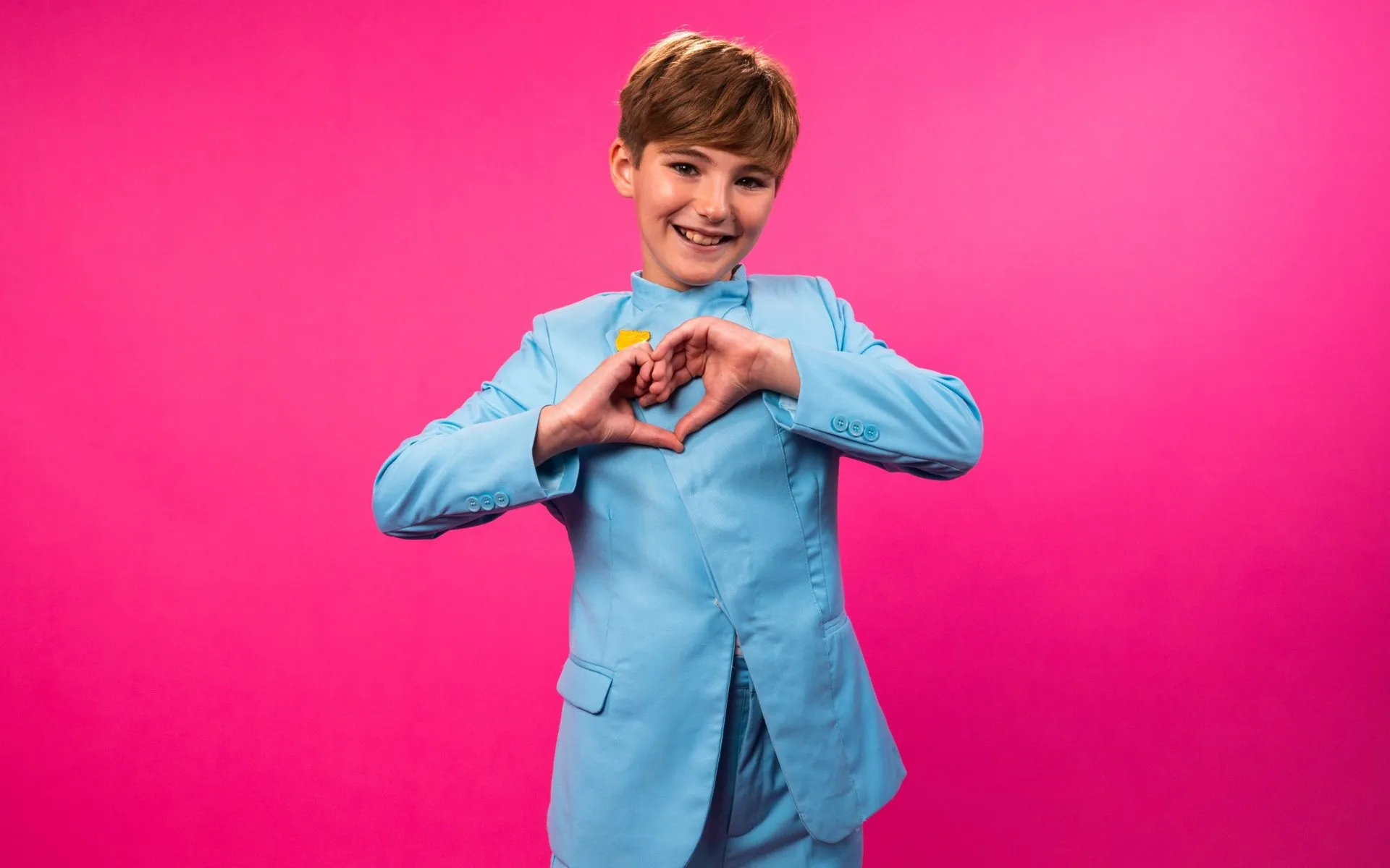 Lucas Paulano, sonriente, hace un corazón con las manos delante de un fondo rosa