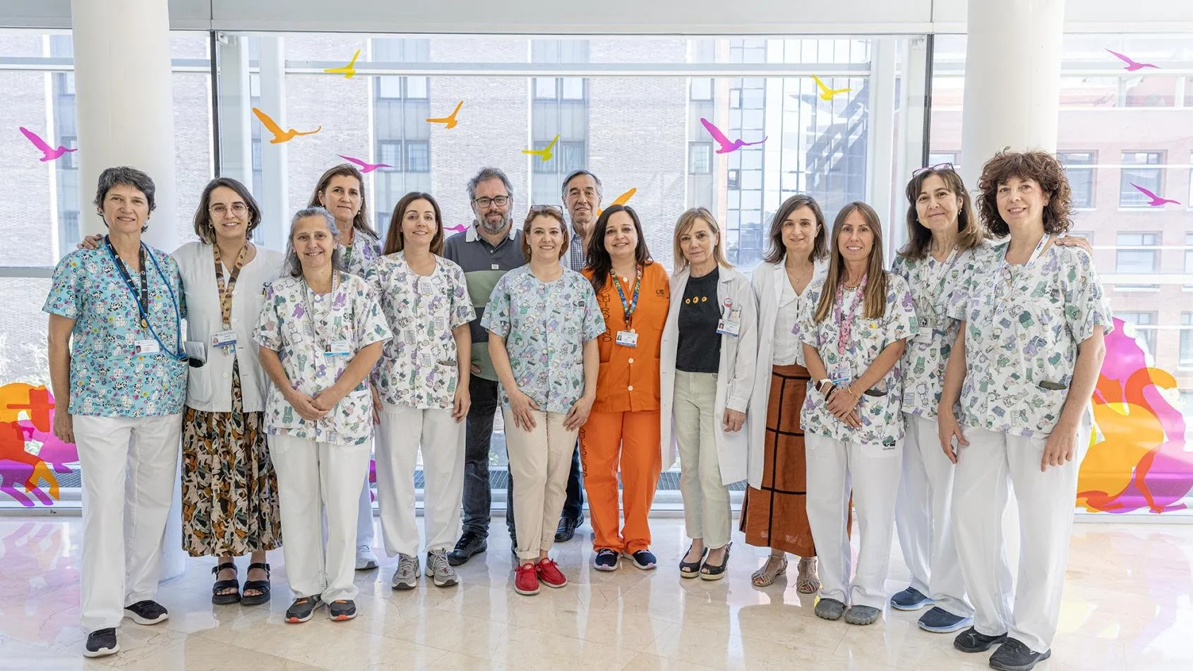 Quince profesionales sanitarios, la mayoría con pijama de estampado infantil o bata blanca, posan juntos en un vestíbulo acristalado decorado con siluetas de pájaros de colores