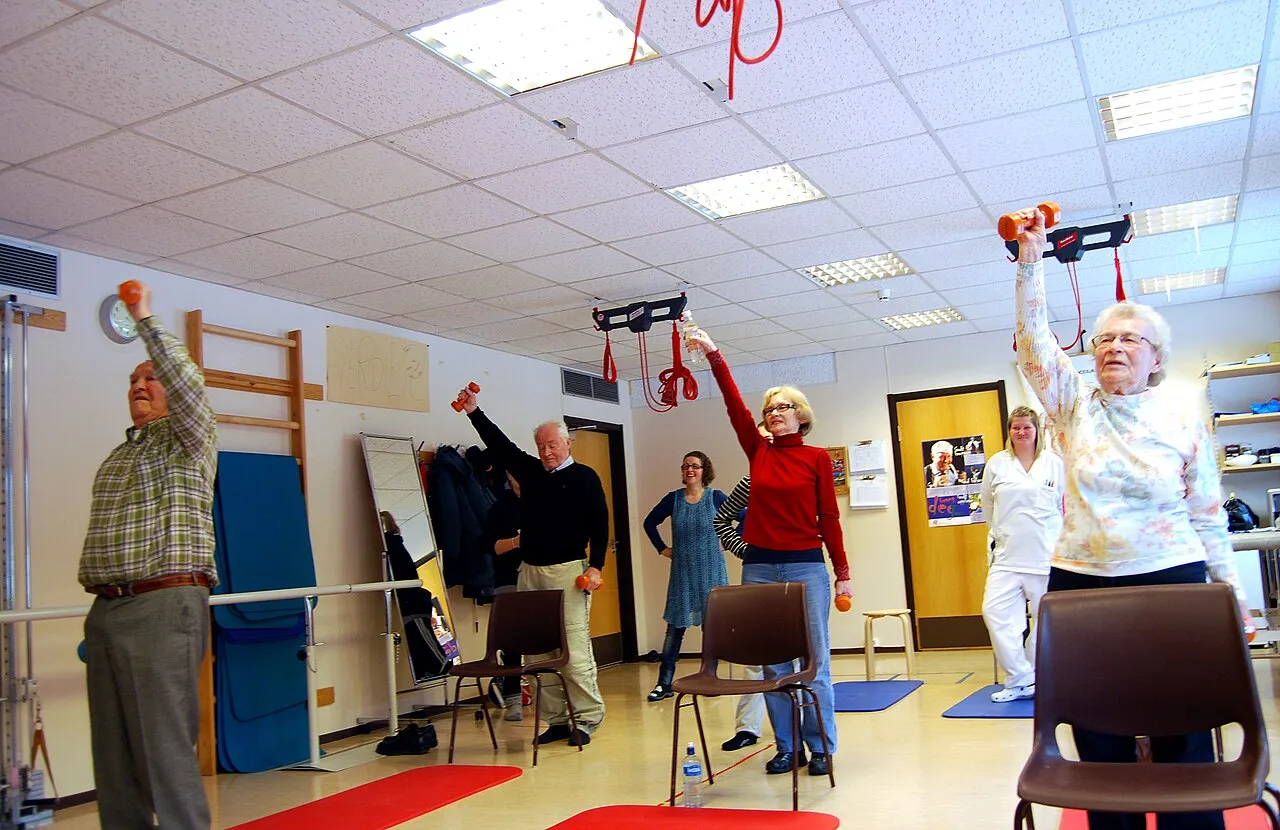 Un grupo de personas mayores levanta pequeñas pesas por encima de la cabeza en una sala de ejercicio