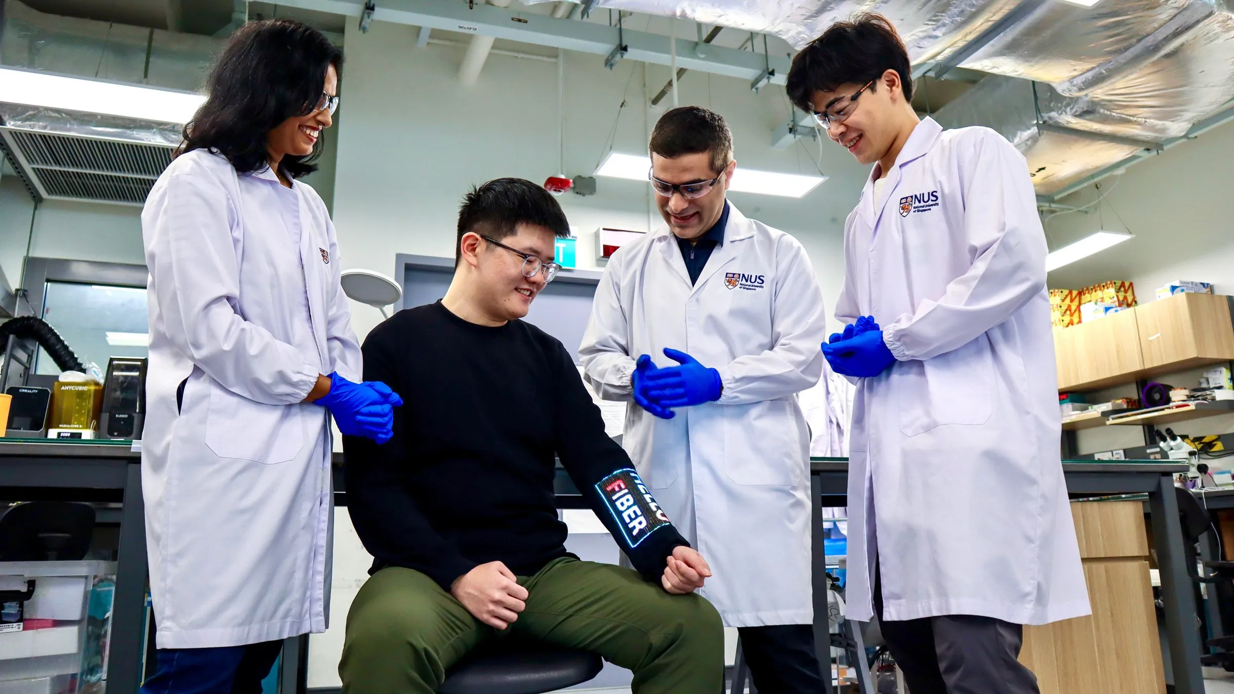 Cuatro investigadores con bata en un laboratorio sonríen mientras uno de ellos, sentado, lleva en el antebrazo la manga con la palabra FIBER iluminada
