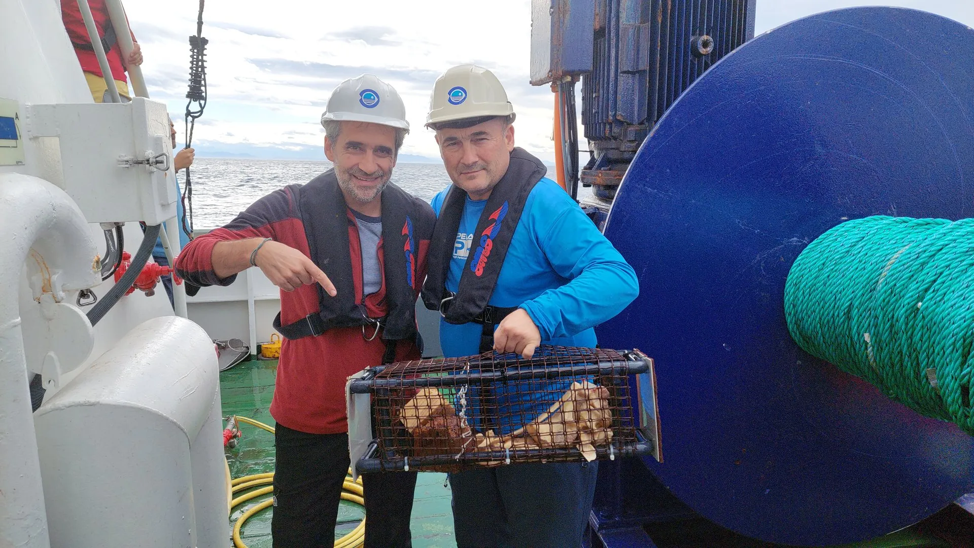 Dos investigadores con casco y chaleco salvavidas sonríen en la cubierta de un barco junto a una cesta metálica llena de huesos