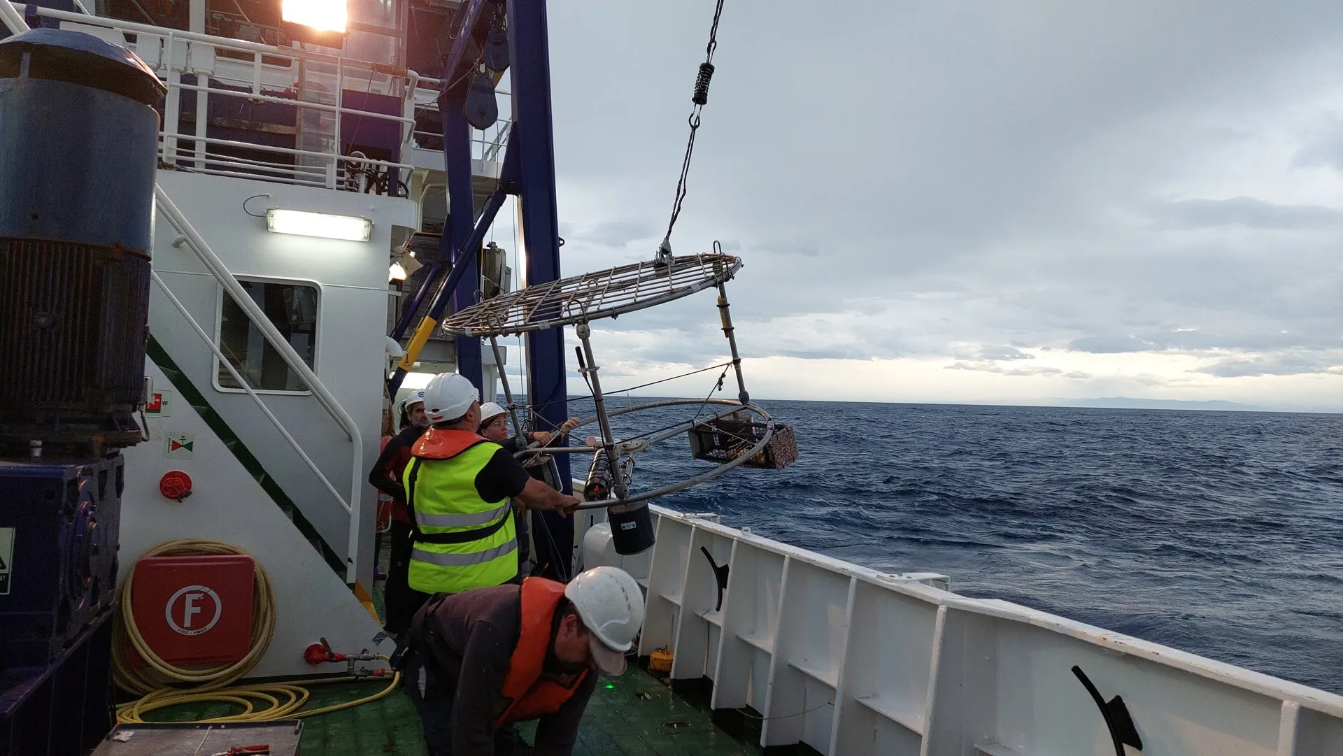 Estructura metálica con flotadores colgada de una grúa sobre la cubierta de un barco, con el mar al fondo