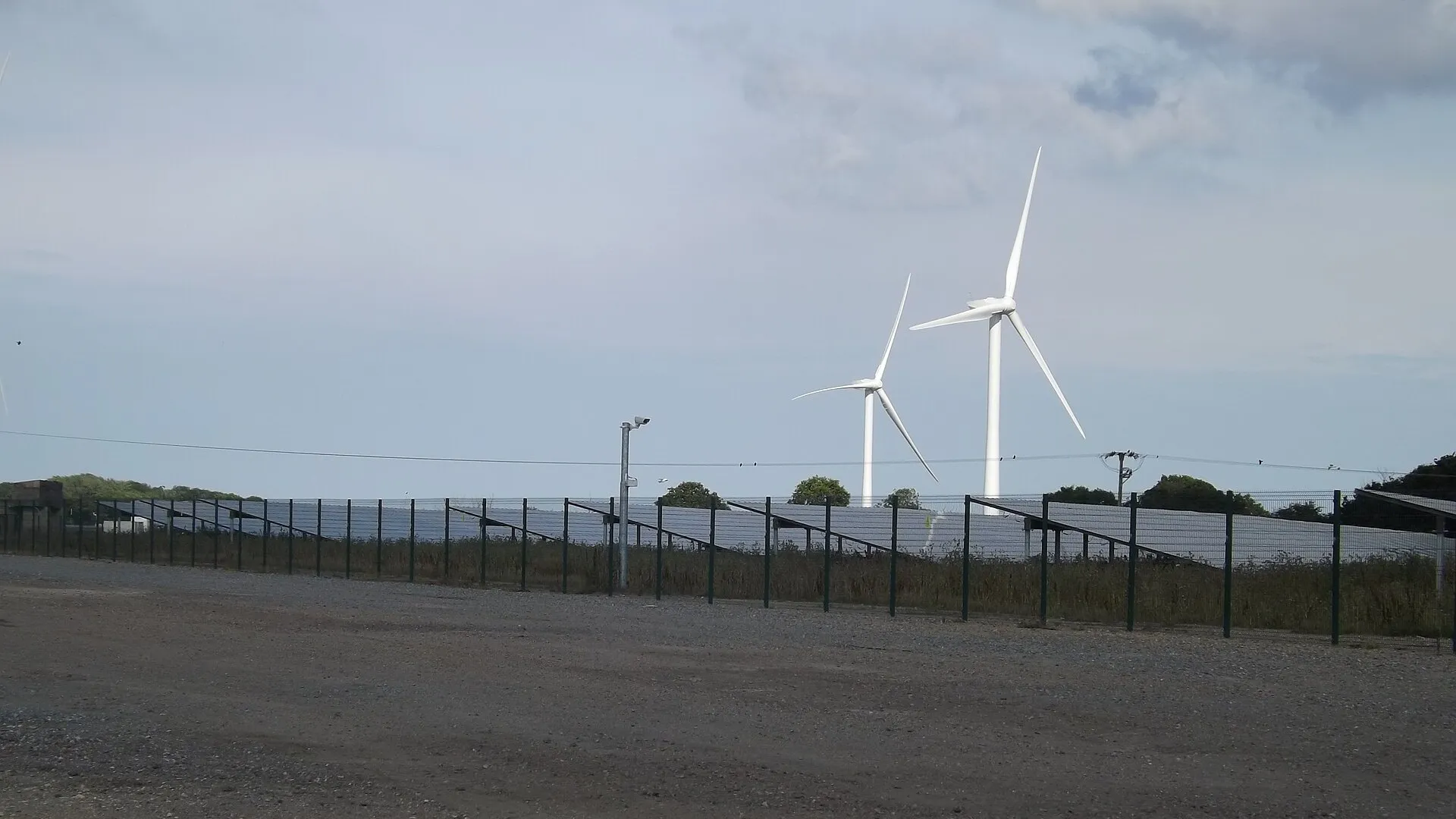 Hileras de paneles solares tras una valla, con dos aerogeneradores blancos al fondo bajo un cielo con nubes