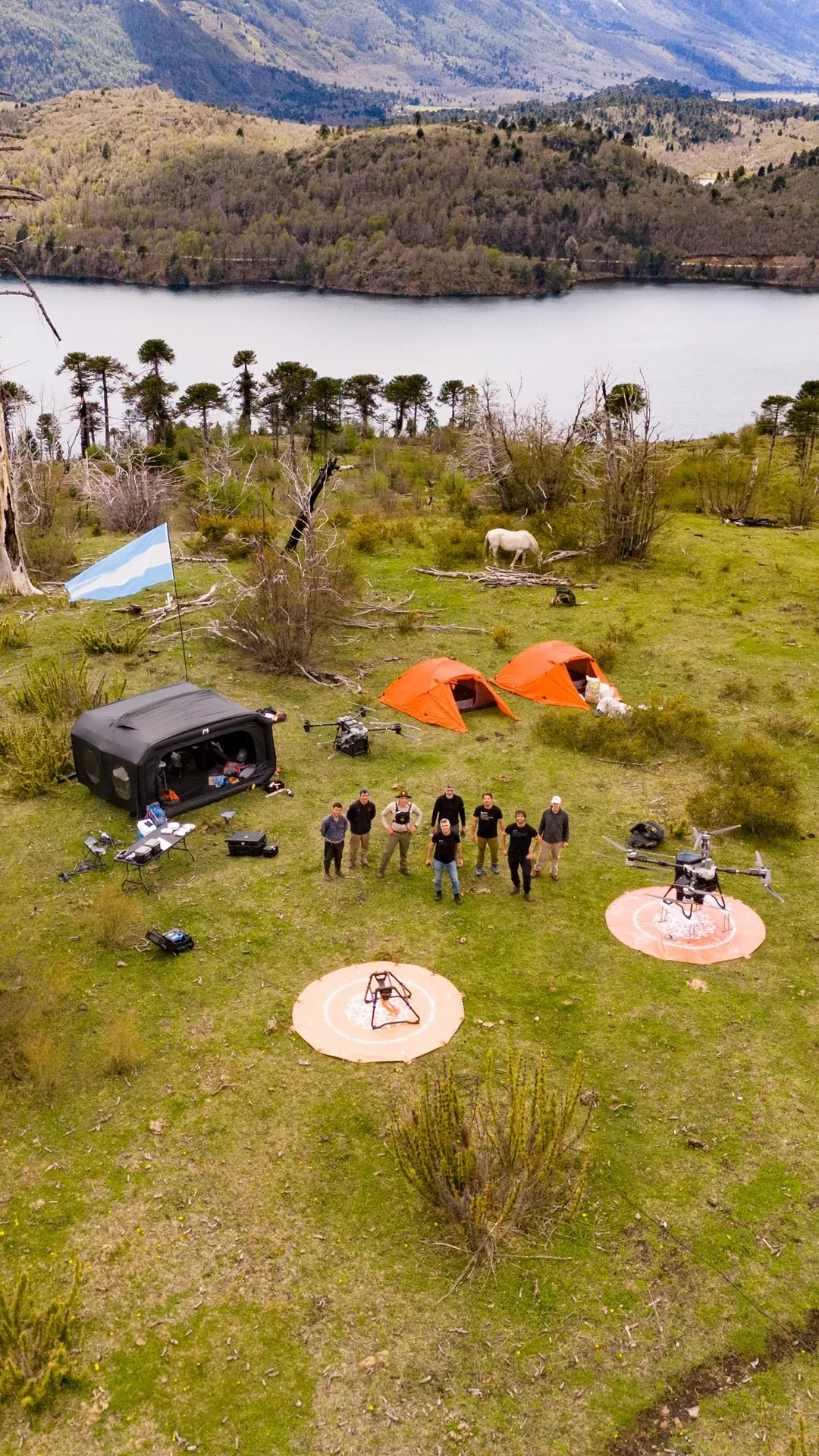 Vista aérea de un campamento en un prado junto a un lago: dos tiendas naranjas, una carpa negra, dos drones sobre plataformas de aterrizaje, un caballo blanco y un grupo de ocho personas mirando a la cámara