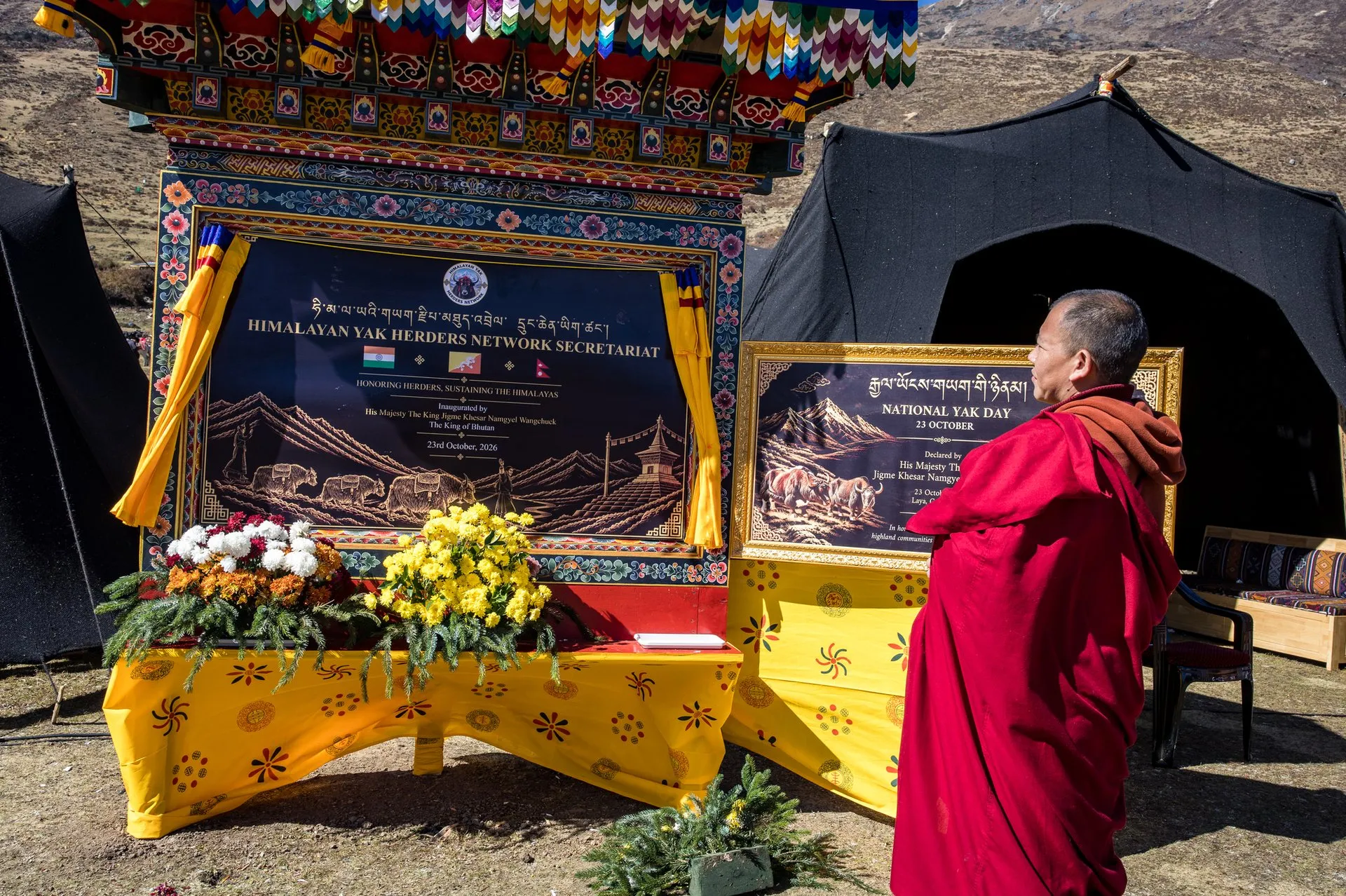 Un monje con túnica roja ante una placa enmarcada en madera pintada en la que se lee «Himalayan Yak Herders Network Secretariat», con las banderas de la India, Bután y Nepal