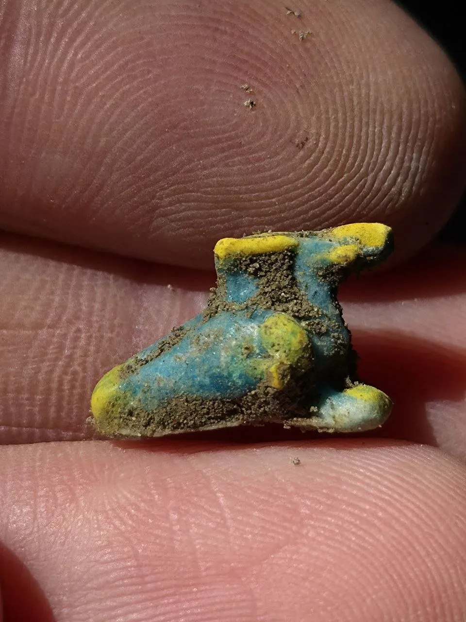 Pequeño amuleto azul y amarillo con forma de sandalia con alas, sostenido entre dos dedos