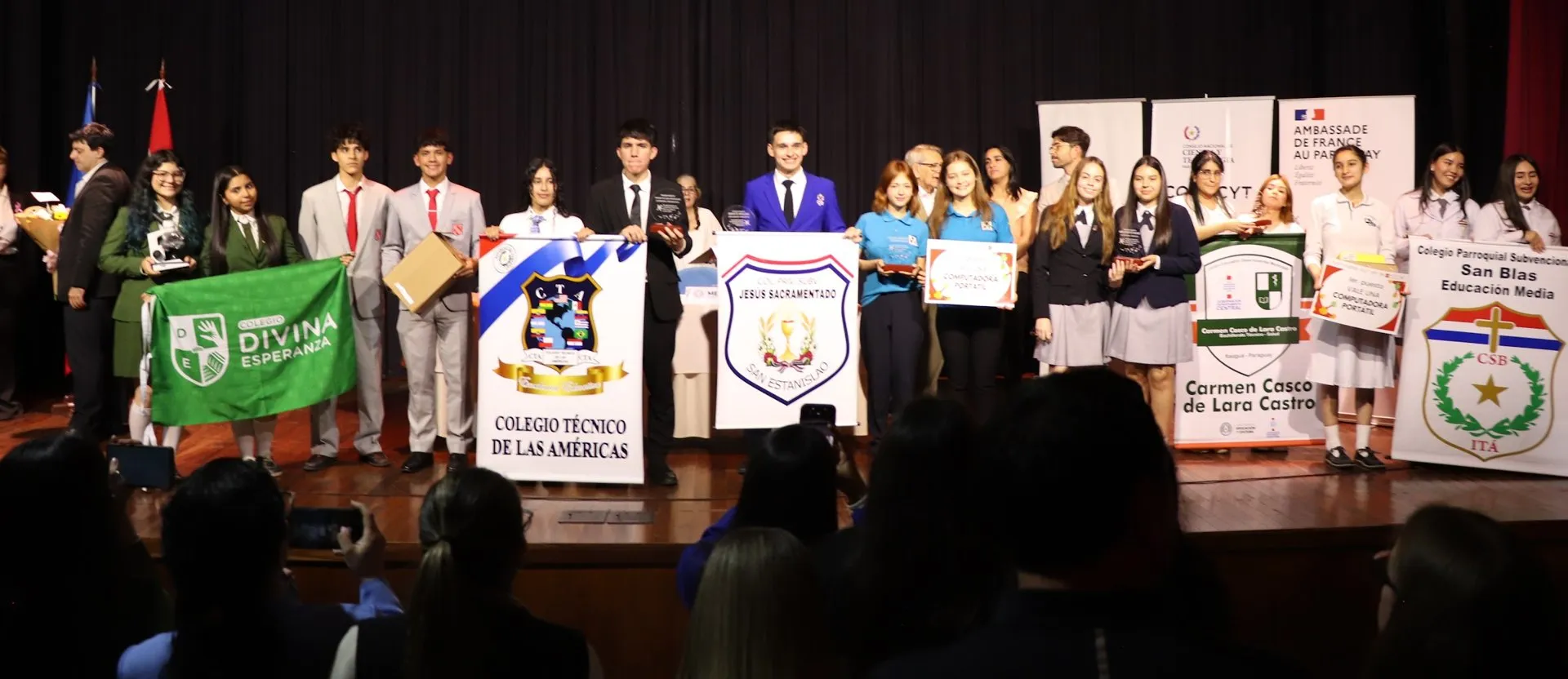 Estudiantes con uniformes de distintos colegios posan en un escenario con los estandartes de sus centros y los premios