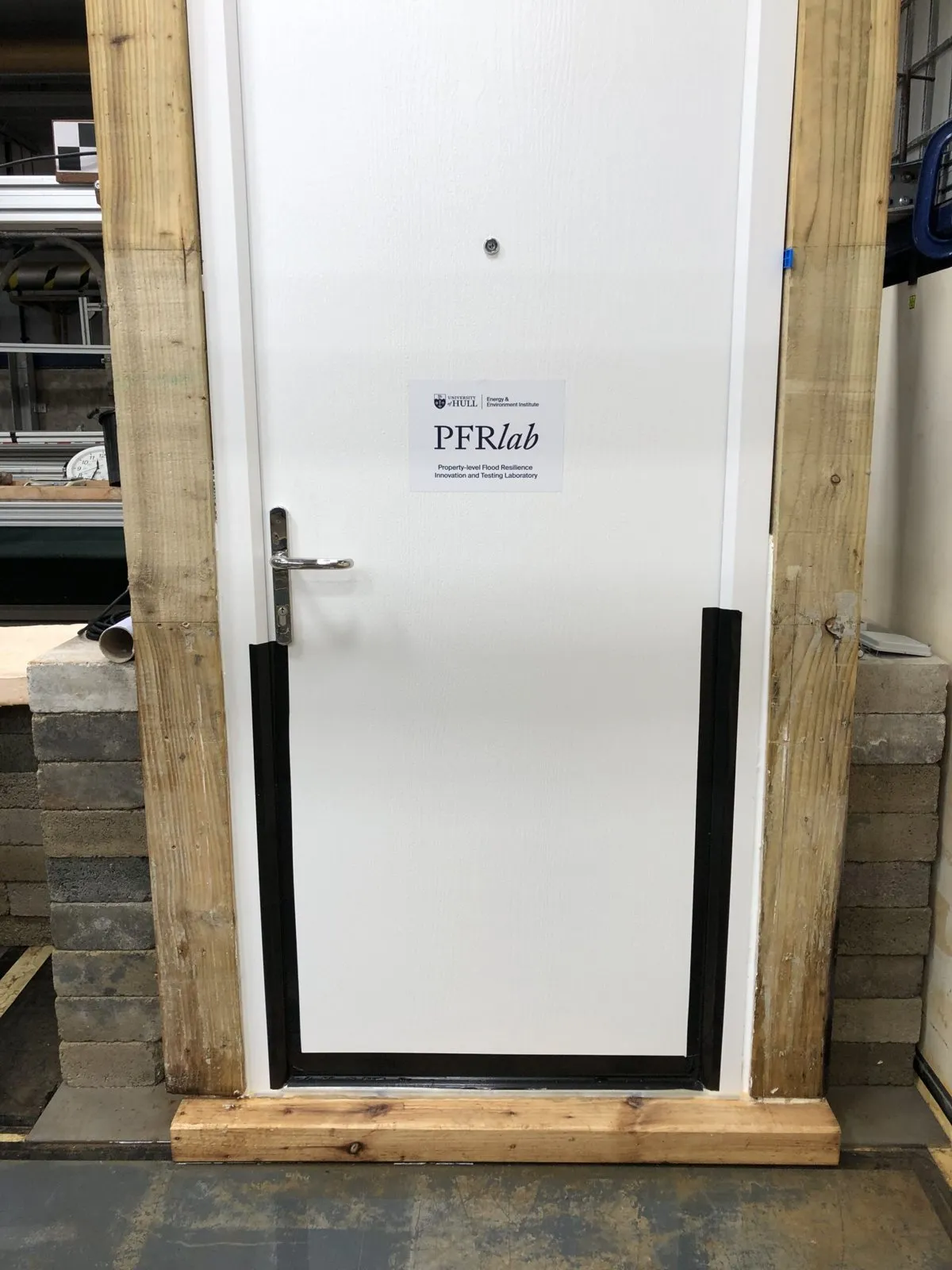 Una puerta de entrada blanca en el laboratorio de inundaciones de la Universidad de Hull, con el borde inferior y los laterales sellados con cinta negra, dentro de un marco de madera de pruebas