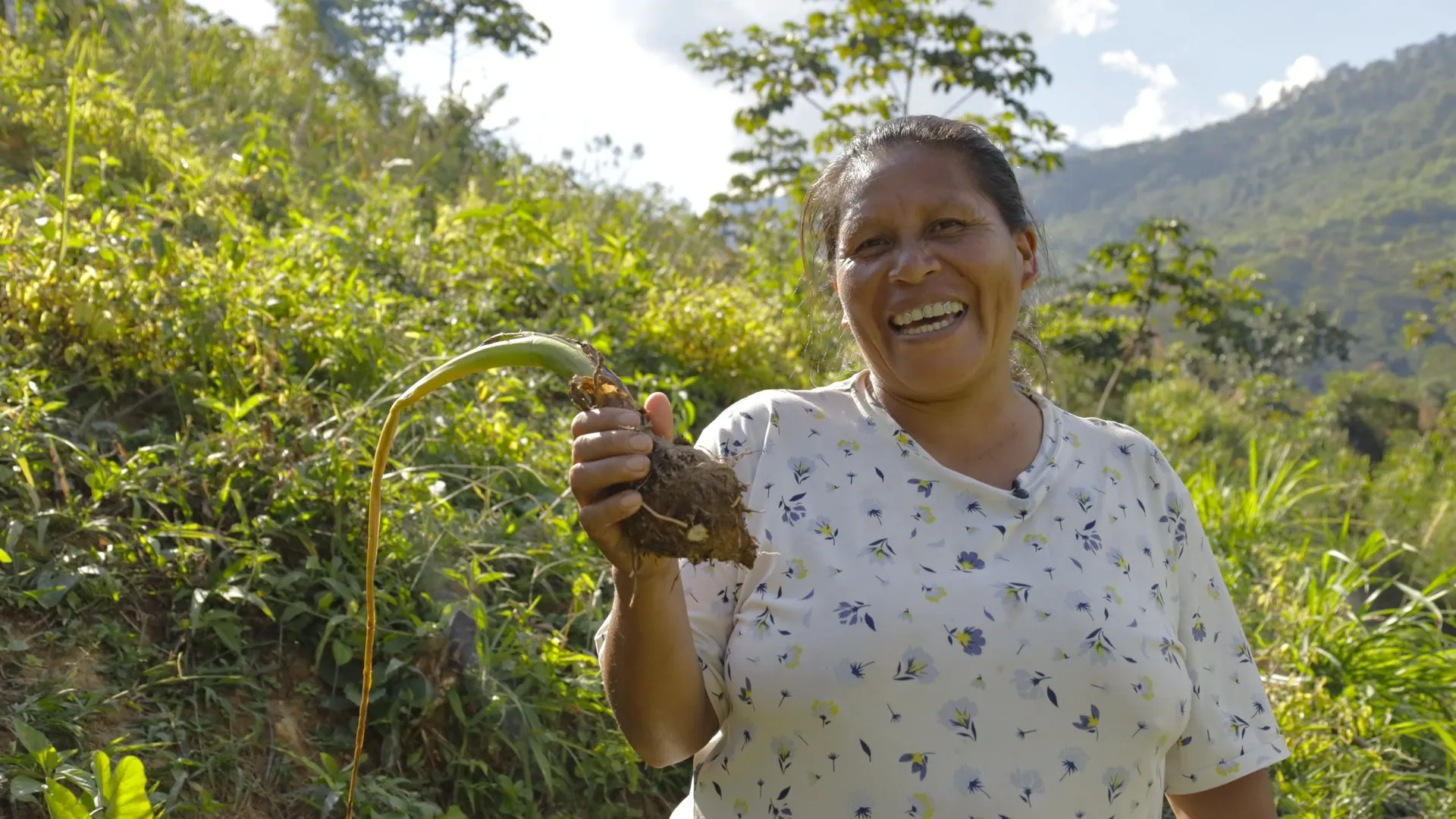 Una mujer sonríe en una ladera verde de la selva con una planta con raíces en la mano y montañas al fondo