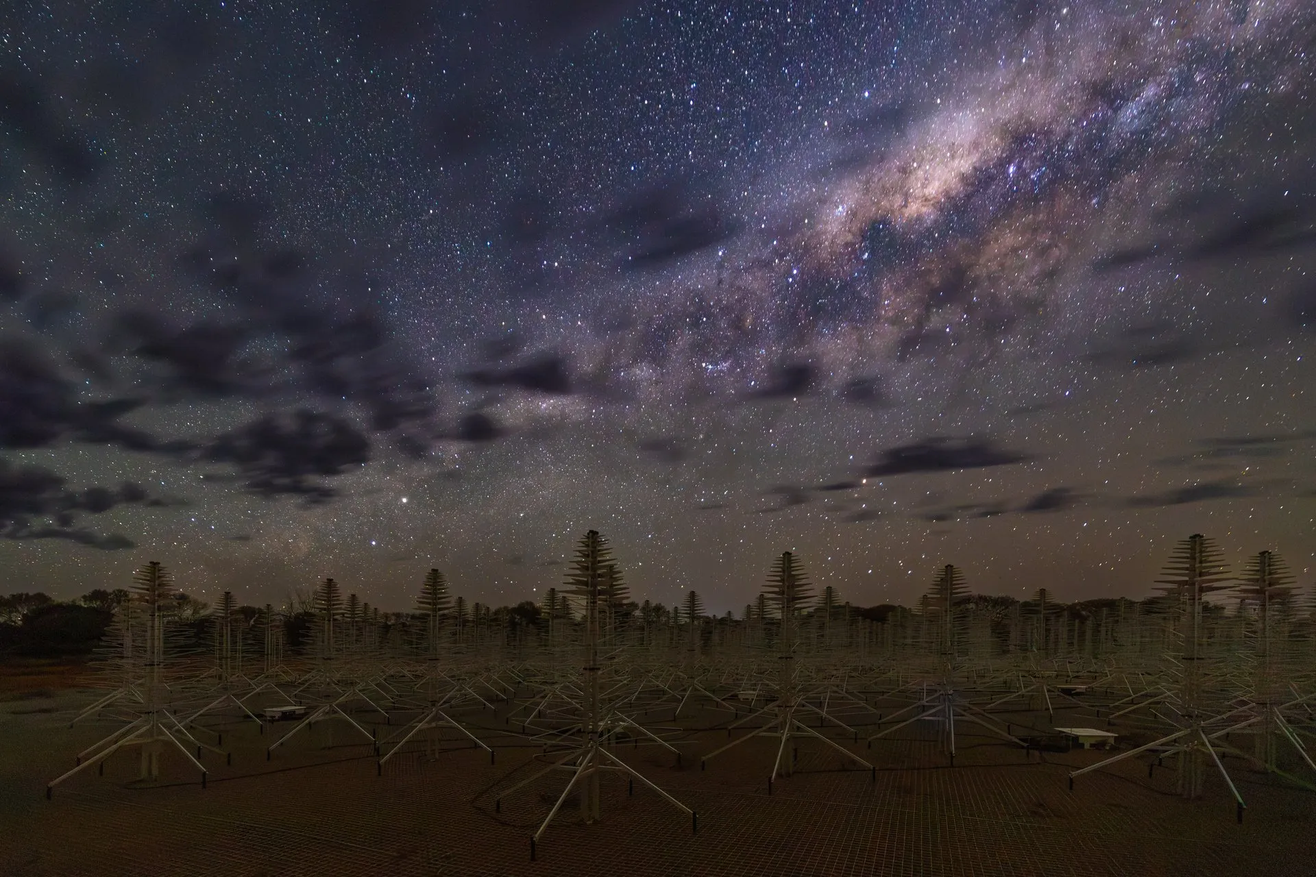 Decenas de antenas metálicas con forma de árbol en un campo rojizo, bajo un cielo nocturno con la Vía Láctea