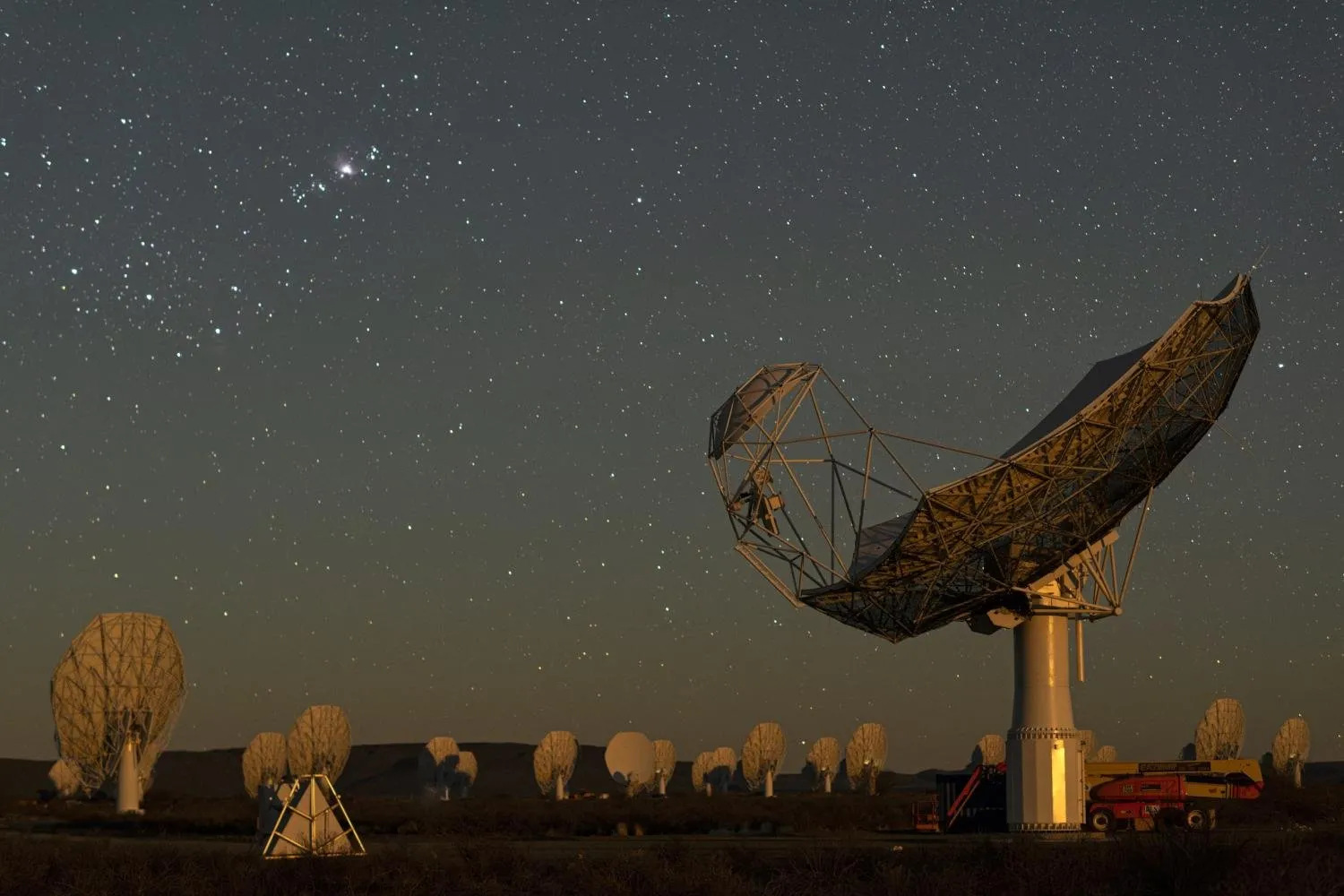 Gran antena parabólica de estructura metálica iluminada, con muchas antenas blancas más pequeñas al fondo bajo un cielo estrellado