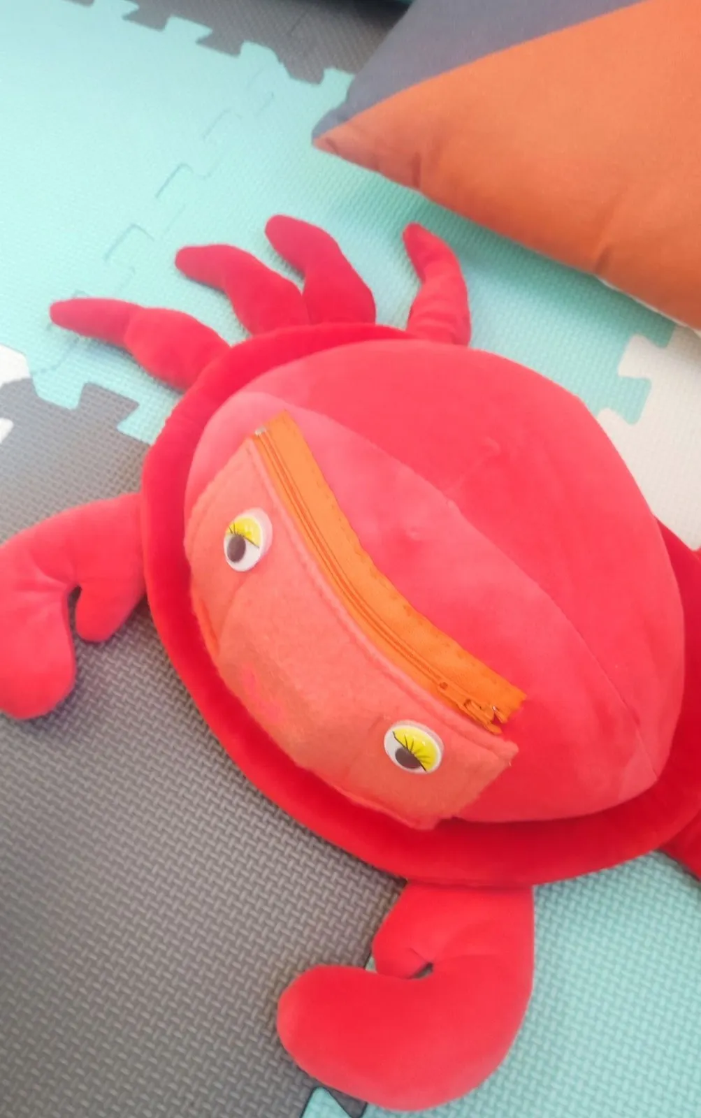 Un cangrejo de peluche rojo, con ojos y una bolsa con cremallera en el caparazón, sobre una alfombra de goma de colores