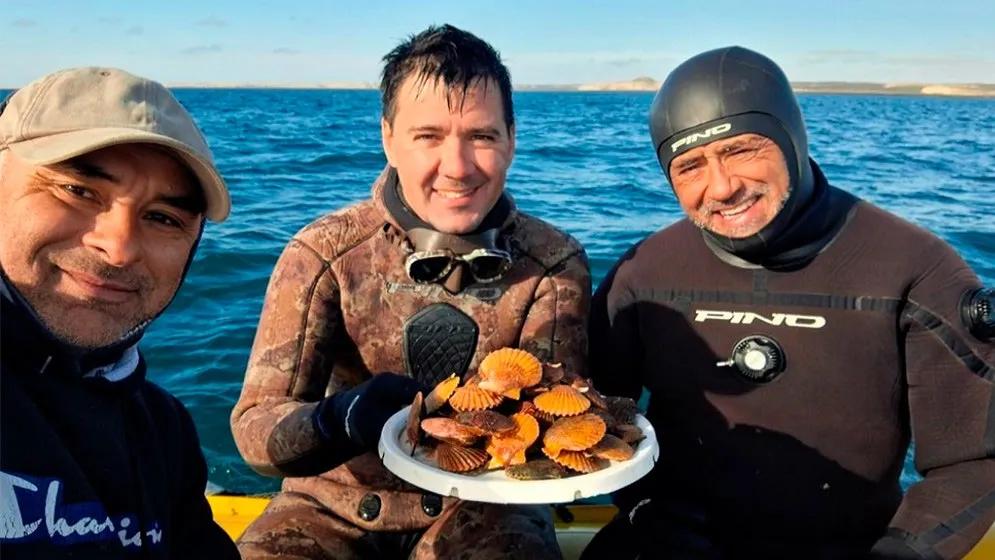 Tres hombres sonrientes en una lancha, dos con traje de buceo, sostienen un plato lleno de vieiras naranjas con el mar y la costa del golfo detrás